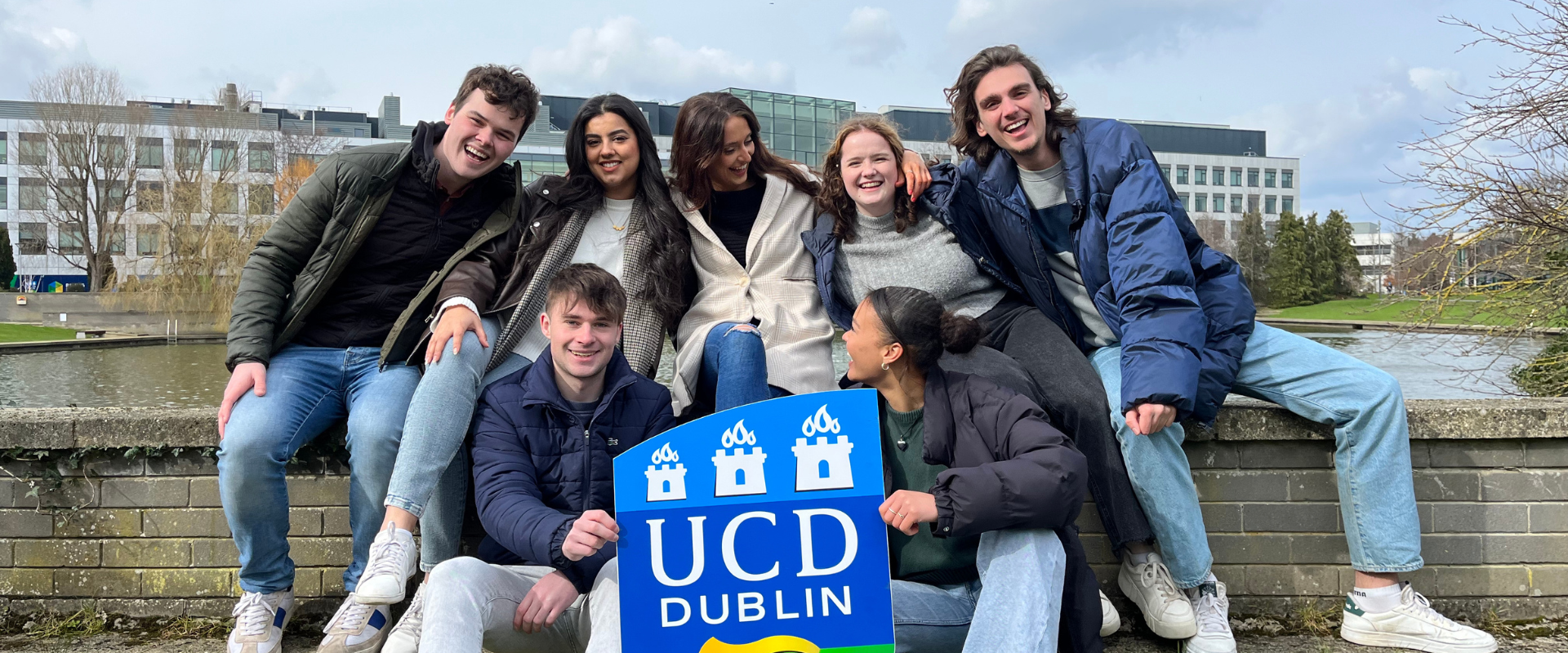 Seven students posing for a photo in front of the Science Building with a UCD logo prop.